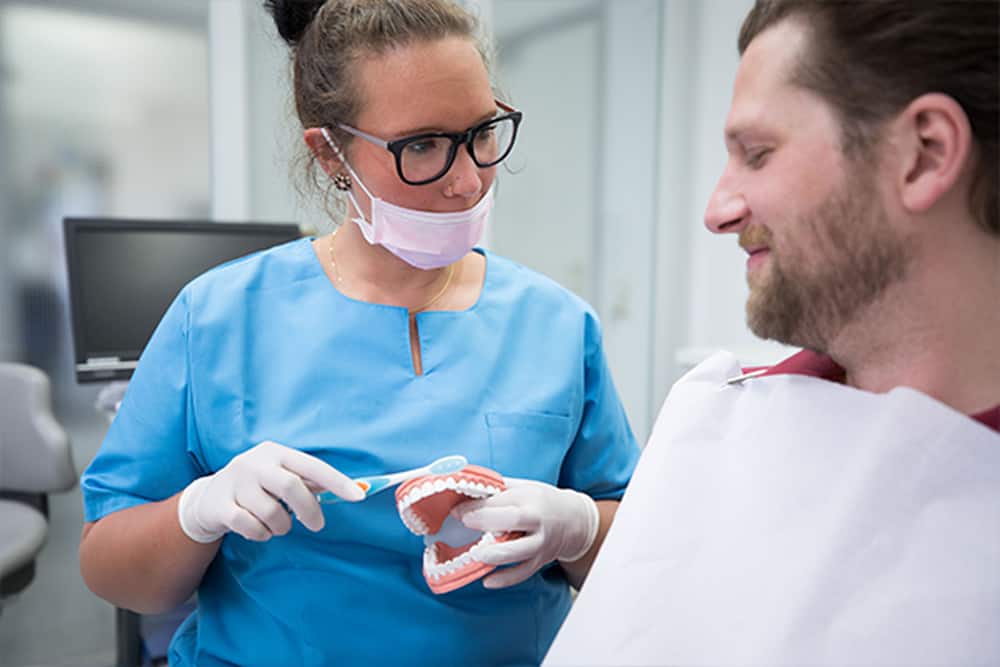 Zahnmedizinische Fachangestellte für die Prophylaxe zeigt einem Patienten die richtige Zahnputztechnik